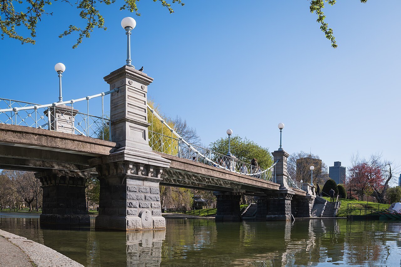 The small suspension bridge across the Public Garden lagoon, between stone piers and iron railings