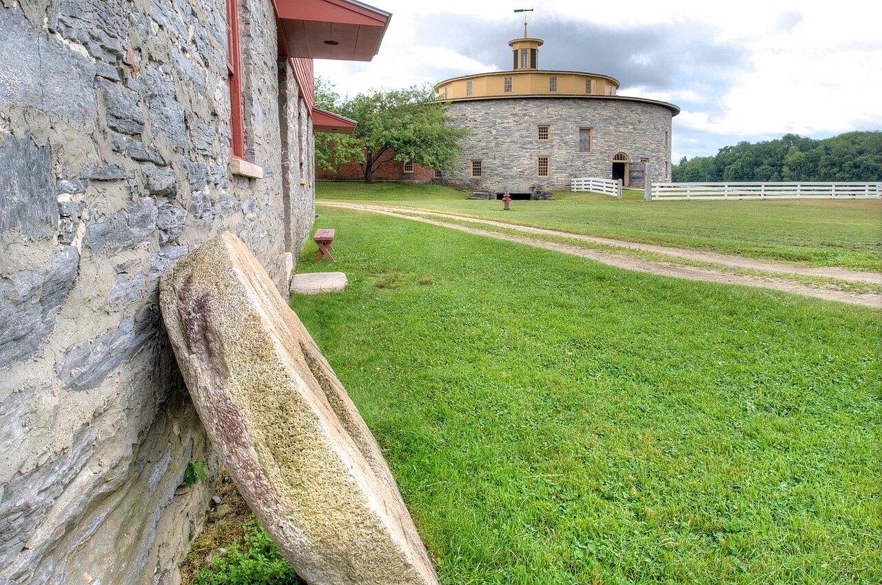 A millstone leaning against a stone wall, the Round Stone Barn across the grass