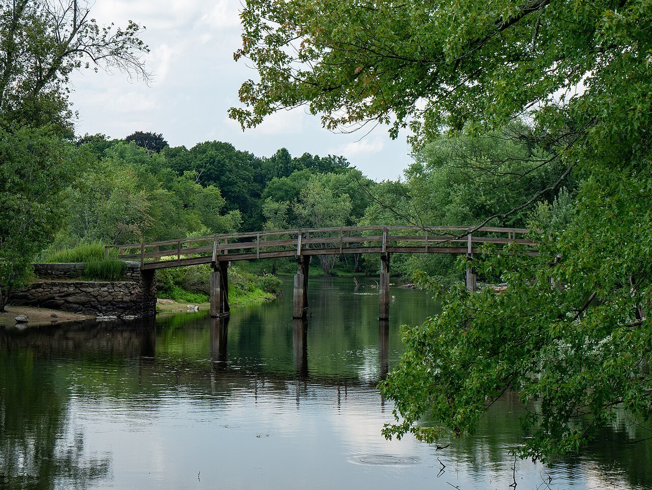 The North Bridge between green trees, reflected in the dark Concord River