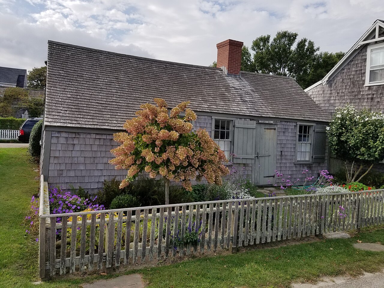 A low shingled cottage in Siasconset behind a picket fence, a flowering shrub in the yard